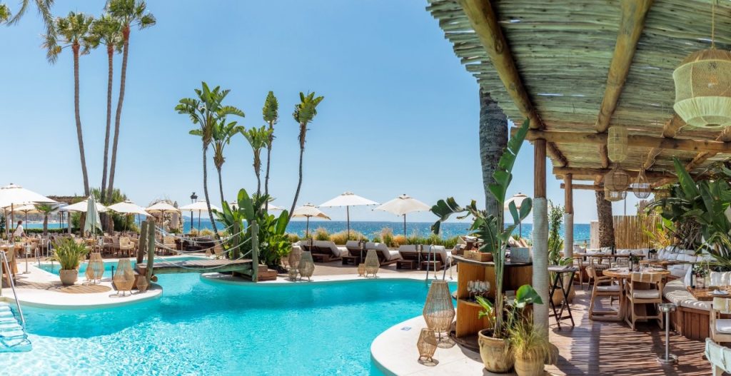 View of Chambao beachclub with a covered timber pergola on teh right, pool in the middle and paraols and a glmipse of the sea in the distance